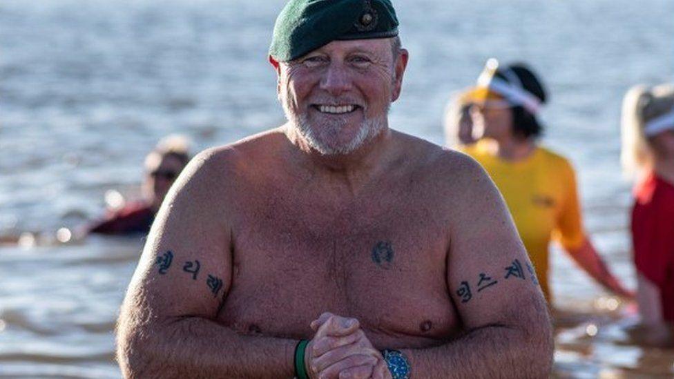 Man standing in sea wearing green beret, he has his hands together in front and tatoos on both upper arms.