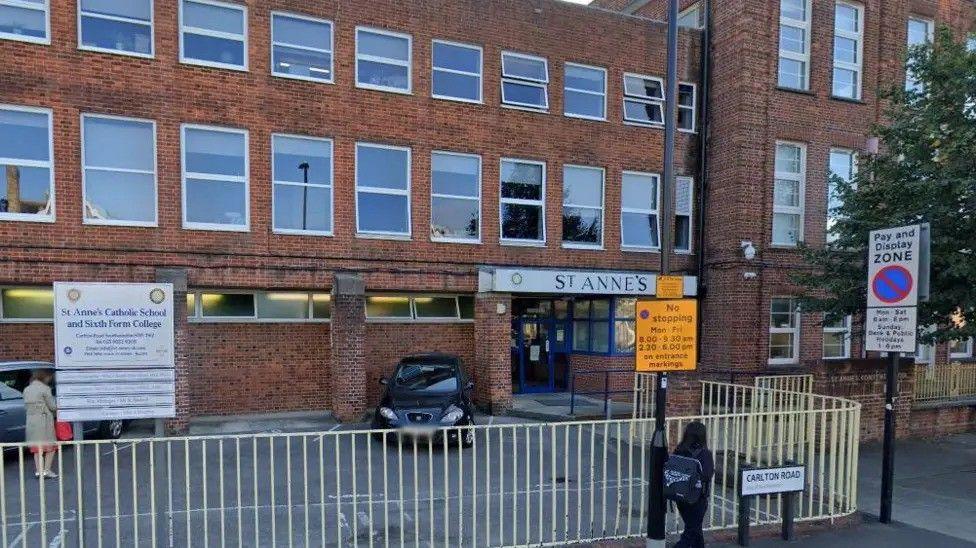 A boxy red bricked building on a main road. A 'St Anne's' sign overhangs a doorway entrance, while a larger school sign stands in the school car park.