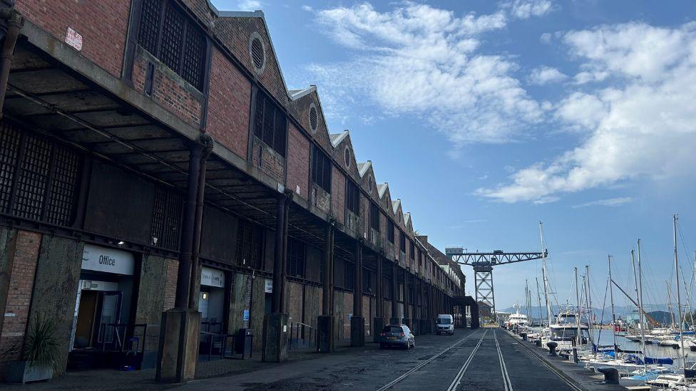 A row of brick warehouse buildings stand in a row on the left hand side of the picture, leading up to a large crane at the end of a tarmacked dockyard area. White sailing yachts in a marina line the right hand side of the dockyard.