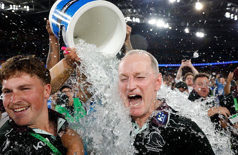Michael Maguire, head coach of the Broncos, celebrates victory following the NRL Grand Final match against the Melbourne Storm in Sydney. Maguire is drenched as water pours from a large blue-and-white cooler over his head, splashing heavily. A cheering crowd and bright stadium lights fill the background.