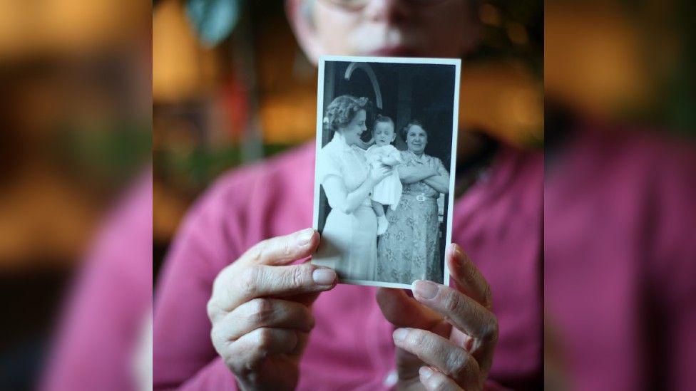 A black and white photo of Adele as a baby being held by her adoptive mother - the photo is being held by an adult Adele who is blurred in the background