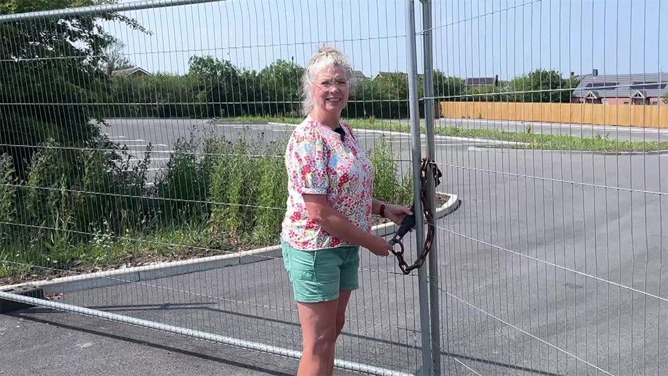 A woman standing in front of a padlocked car park. She has blonde hair and is wearing glasses, a floral top and green shorts.