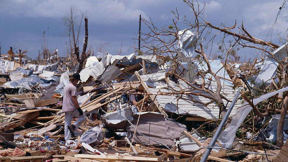 A man surveys the wreckage of buildings and trees with a mix of blue sky and clouds overhead