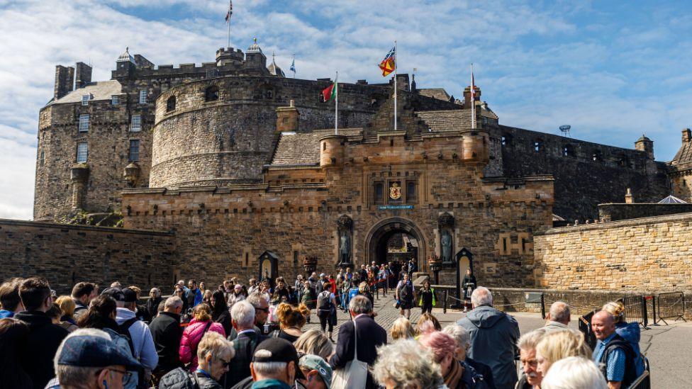 A crowd of people filing into a large medieval-style castle. Steep stone walls, turrets and towers rise from a flat entrance flanked by smaller walls. Colourful flags flutter against a blue sky. 