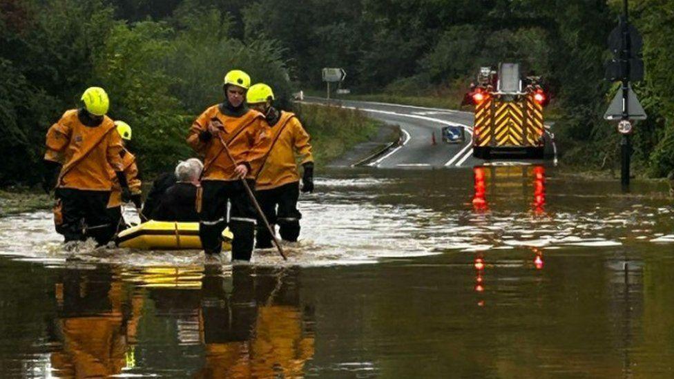 A fire engine is seen at the edge of a heavily flooded area with fire crews in the foreground pulling a yellow boat with people onboard through the water