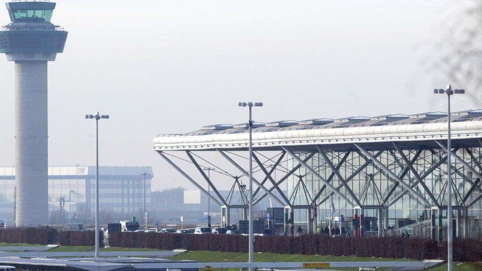 Stansted Airport, showing the tall control tower and white roof of the terminal building. Cars are arriving at the drop-off at the terminal, with people entering the building.