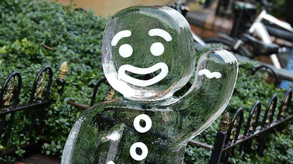 A ice sculpture of a waving gingerbread man stands in front of black railing and a hedge