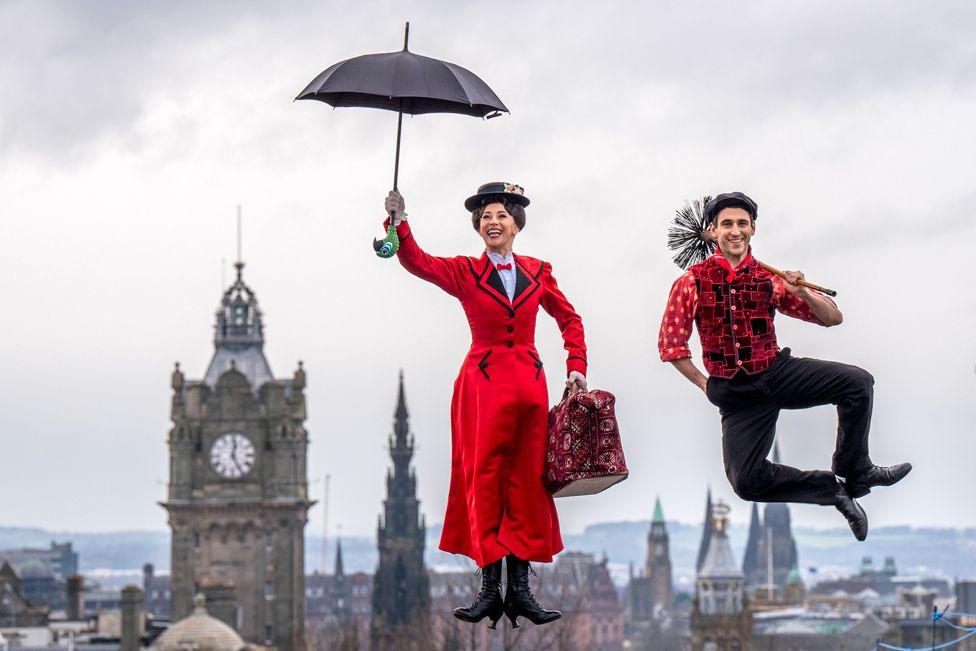 Stefanie Jones, dressed as Mary Poppins - in a red coat and holding a red umbrella -  and Jack Chambers, dressed as Bert -  in black trousers, red shirt and waistcoat and a black flat cap, holding a sweep - pose by jumping in the air, during a photo call on Calton Hill, with the Edinburgh skyline in the background.