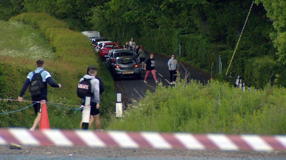Cars parked in a narrow country road with large numbers of people walking along the road. Red and white tape can be seen in the foreground