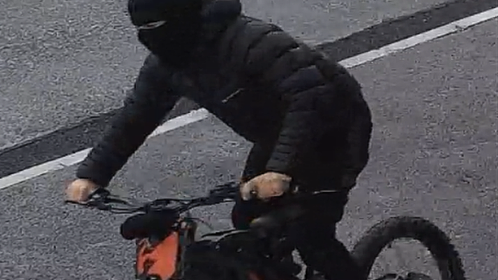 A man dressed all in black rides an electric bike with an orange panel under the handle-bars. He is wearing a  balaclava, a bubble coat and tracksuit bottoms. 