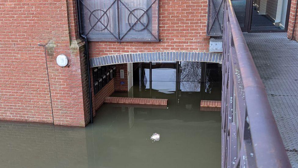 A shot looking down on to flood waters surrounding a red brick, relatively modern-looking building. The dark green water is several bricks high and can be seen reaching underneath an arched area, through some doors and into a lobby, which appears to also be underwater. A row of mail boxes are just visible underneath the arch and are around six bricks above the water. A raised metal pathway and handrail can be seen on the right of the image. 