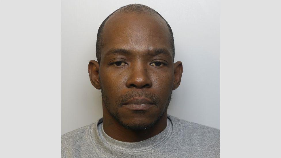 A police mugshot of David Malcolm. He is wearing a grey t-shirt and grey jumper, and standing against a light grey background, looking at the camera. 