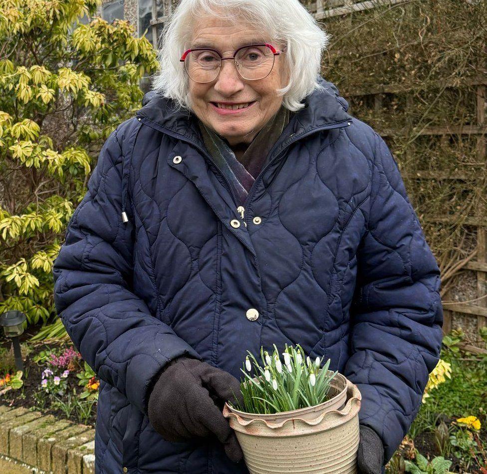 Maureen is wearing a thick padded blue jacket. She has white hair and glasses and is holding a flower pot with snowdrops in it. She has thick black gloves on.