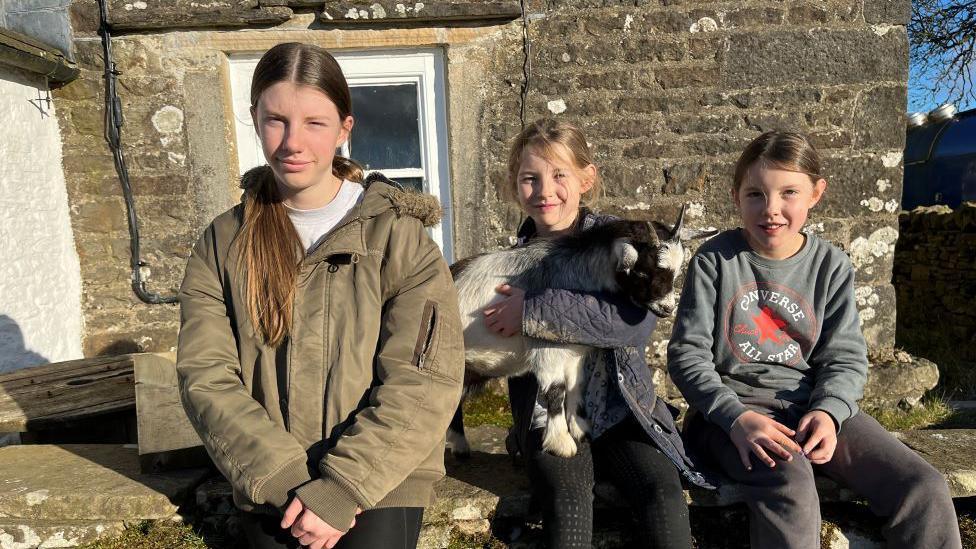 Three girls wearing outdoor clothing are sitting on a wall outside a stone house. The middle girl is holding a small black and white goat.