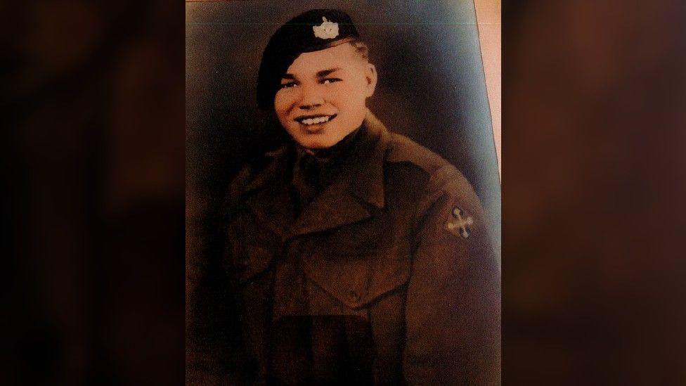 Roy Mills as a young man in military clothing, with a cap, smiling at the camera in a portrait-style photo.