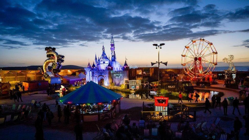 An aerial image taken at dusk showing the illuminated art installations at Dismaland. There is a large Disney castle lit up with blue and purple lights in the centre, which is reflecting on the water in the abandoned lido below. There is a large red Ferris wheel on the right, and a Punch and Judy stall in the foreground. The sky is cloudy and blue with an orange hue on the horizon.