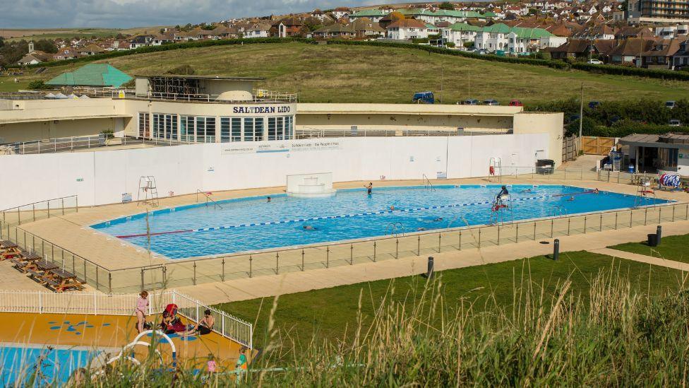 A swimming pool located next to an area of grass. People are sat around by the pool in Saltdean. Houses can be seen in the background. 