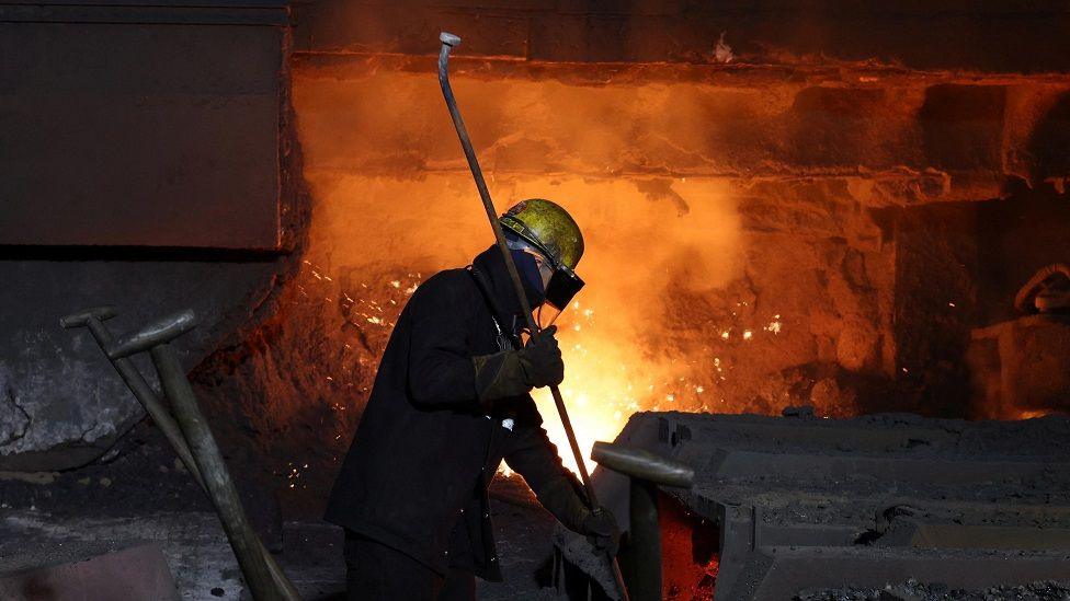 A worker in black protective wear and a grubby yellow safety hat stokes a glowing furnace with a long metal pole at British Steel's steelworks site in Scunthorpe