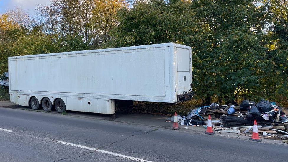 The side view of a white lorry trailer. On its right is fly-tipped waste, including black plastic bags full of waste, wood offcuts and other waste which have been coned of with orange and white cones. They are on a lay-by beside a road