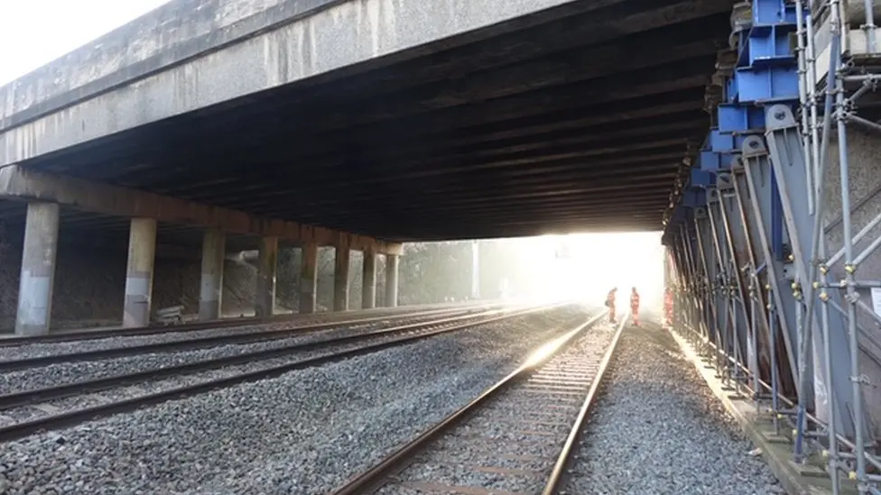 A picture of the Kennington Bridge, with three workmen in orange coats and trousers stood besides one of the three railway tracks that run underneath it 