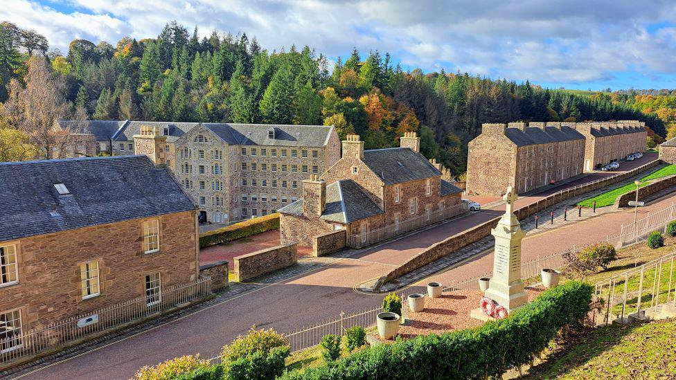 Brown and grey stone buildings. There is a road running along in front of them and there is also a memorial in the foreground.