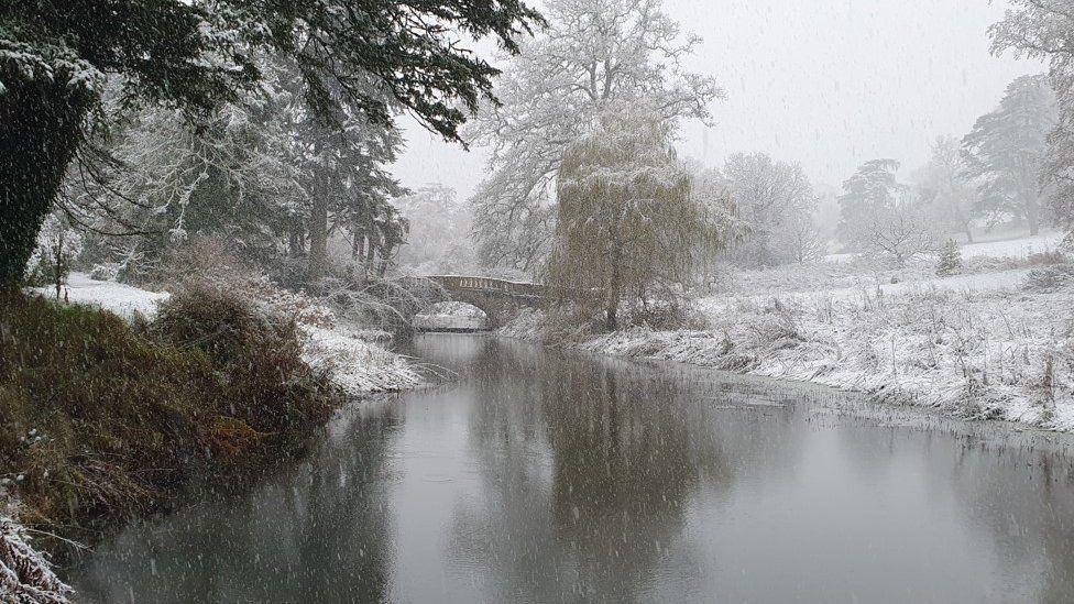 Snow falling on water with icy banks either side and a arched bridge in the distance.