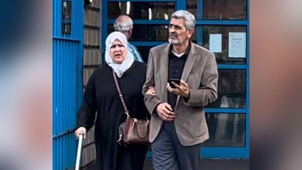 A woman holding a walking stick links arms with a man wearing a suit as they leave a court building, with the man holding a phone in his hand