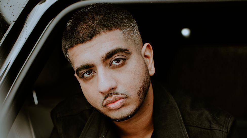 A man sitting in a car, looking outside with a serious expression. he is wearing a black jacket.