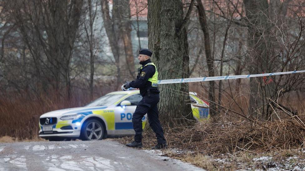 An officer unrolls police tape over a road which is icy, with a police car in the background.