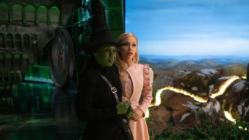 Elphaba dressed in a black hat and dress has a smirk on her face whilst Glinda, dressed all in pink, appears surprised.