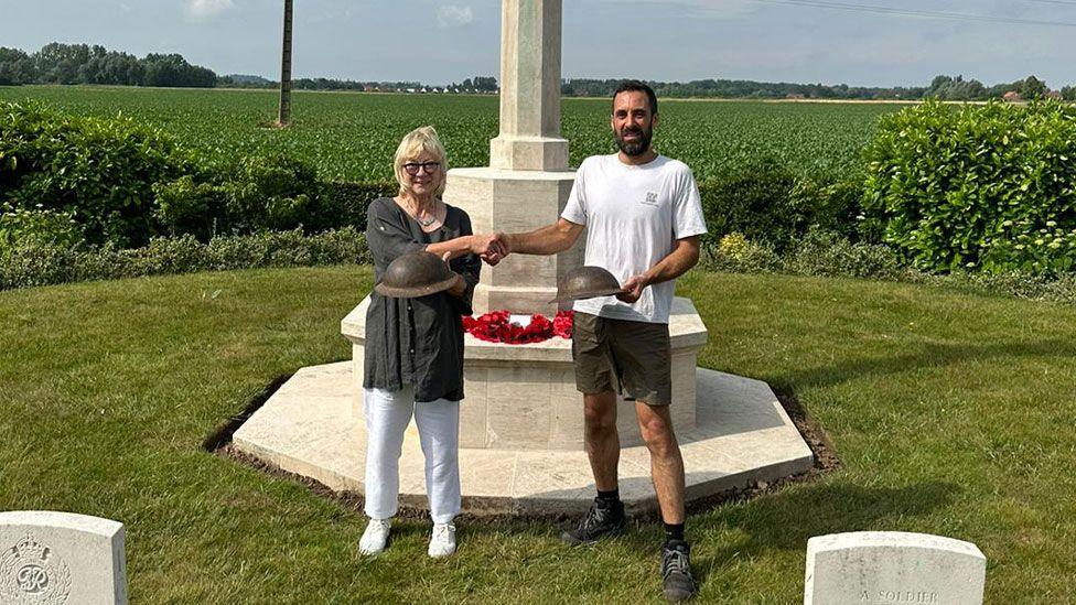 Biddy Fisher wearing a grey top and white trousers shaking hands with Jean-Charles Dufour at the white stone Le Paradis war memorial. She is holding one helmet while he is holding another. Behind them are green fields and sky.