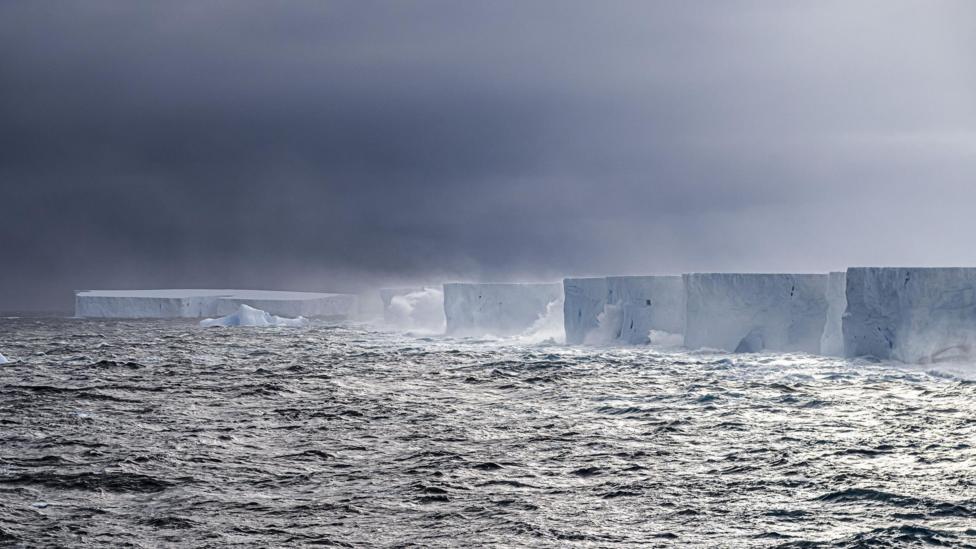 Ice cliffs are lashed by waves in a stormy sea against a grey sky