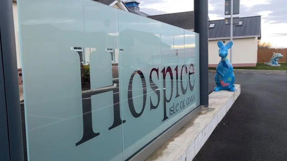 A glass sign that reads Hospice Isle of Man. A blue decorated statue of a wallaby can be seen next to the sign and a similarly painted dolphin statue in in the background.