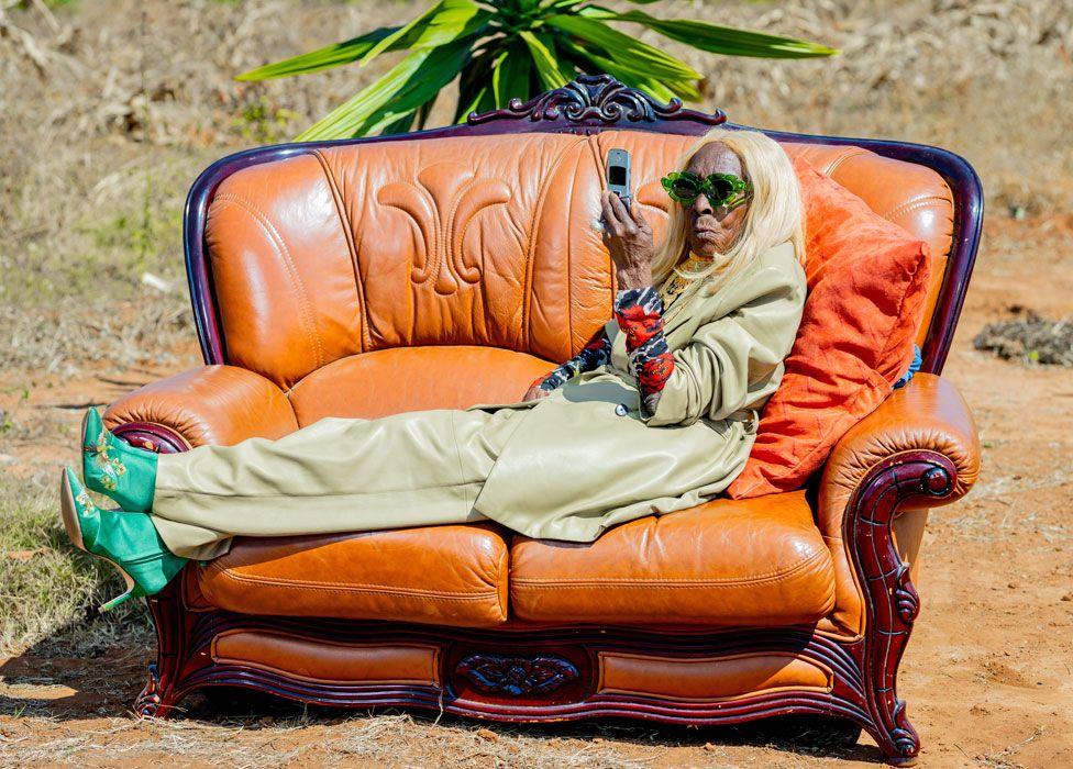 Margret Chola lying on a orange leather sofa in a garden looking at her phone. She has on a wig of blond hair and is wearing a pale green leather loose-fitting trouser suit with green high-heeled boots and green-rimmed sunglasses 