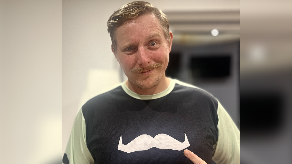 A man with a blonde moustache and hair looking at the camera, he is pointing at his shirt which has a white moustache on it