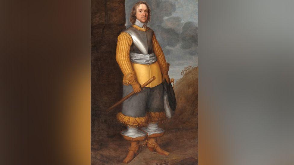 A portrait of Oliver Cromwell, who has shoulder-length born hair, a fine lace collar done up with tassels under his neck, wearing a tawny orange/yellow doublet under a short section of body armour and a light grey silk scarf around his middle. He is wearing grey trousers which end under his knee in ruched orange material. His stockings are white and his tan-coloured boots have very wide cuffs, lined with white material. He is wearing leather gloves with fur trim and holidng a hat in his left hand and a staff in his right hand.