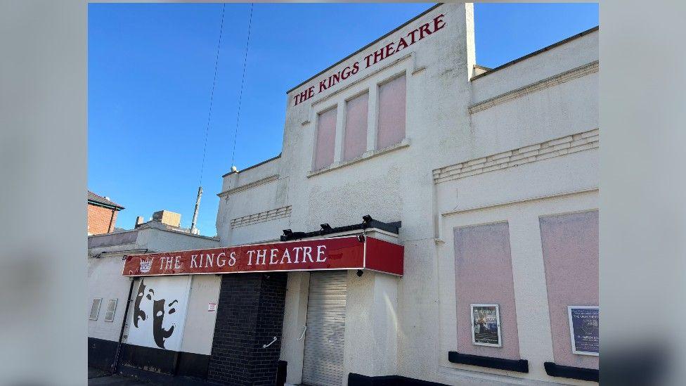 A community theatre under a cloudless blue sky in Gloucester. It is a podium-shaped building painted white with comedy and drama masks painted on the front. It has two red signs saying "The Kings Theatre", and there are some small posters on the front, advertising shows.