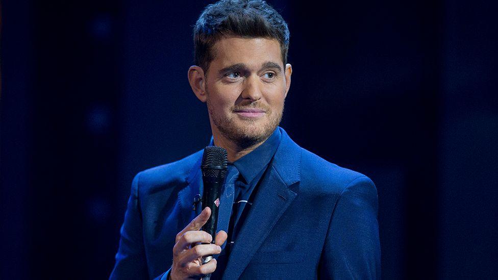Michael Bublé stands on a dark stage in a royal blue suit, shirt and tie. He is holding the microphone in his right hand, looking to the left nd smiling slightly. He has thick brown hair and stubble.