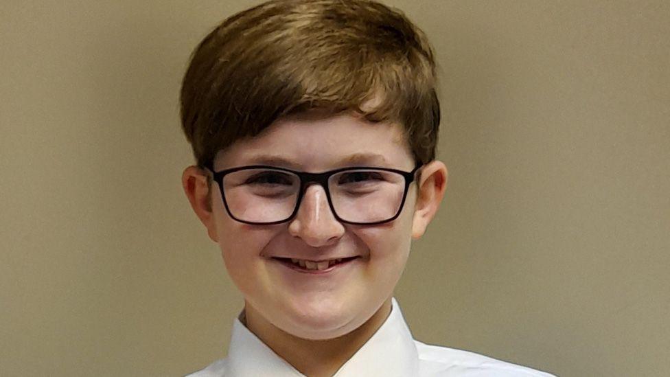 Daniel Flemen before the brain tumour affected his health. He has short brown hair, black-framed glasses, is wearing a white shirt and is smiling in front of a plain beige wall.