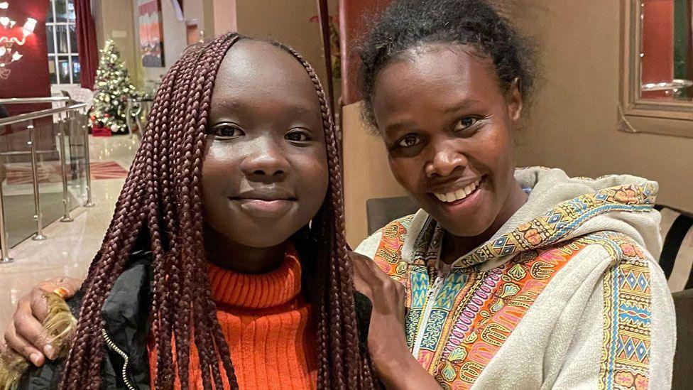 Milcah Cherotich poses in a hotel lobby in London with her hands on the shoulders of Michelle Lumuya Ikeny, who is wearing long braids and an orange jumper