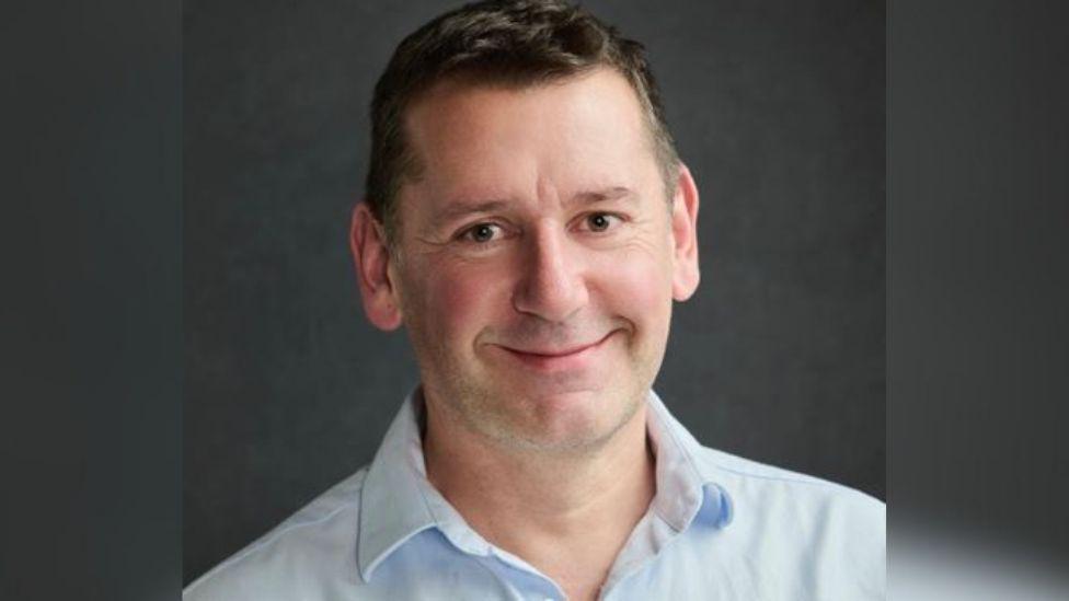 A man looks at the camera wearing a blue shirt.