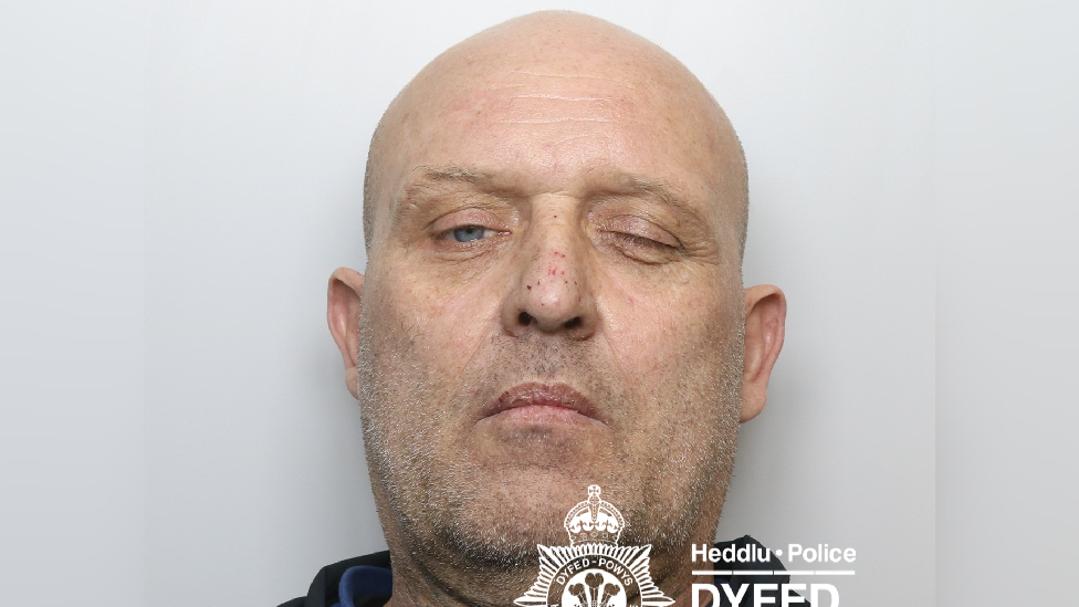Police issue custodu photograph of a 51 year old man with a bald head and greying stubble. His left eye is closed and he's stood in front of a white backdrop.