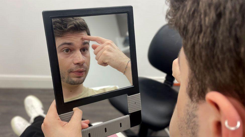 Ven Grecu is looking in the mirror. He is a 24 year old man with brown hair and a faint beard. He is pointing at his forehead in the mirror.