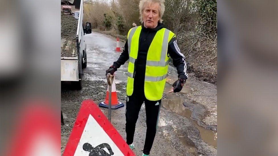 Sir Rod Stewart is standing on a road which has potholes filled with rain water. He is holding a shovel and is dressed in a black tracksuit and a yellow hi-vis jacket. Behind him is a lorry, emptying gravel onto the road