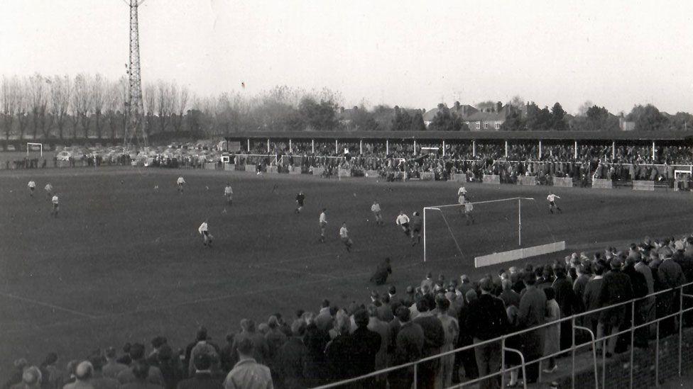 A black and white image of a football match taking place at Cambridge City's Milton Road ground. Men are scattered across the pitch and crowds of people can be seen standing and watching the match along the bottom and top of the ground. The people along the top of the ground are part-covered by a roof