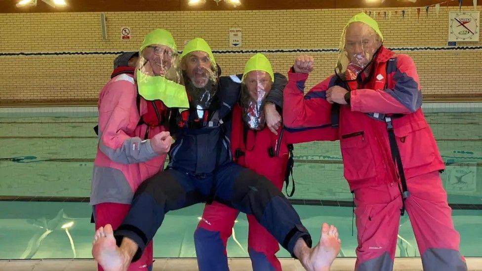 The same four men as pictured in the first image. Here they are stood at the side of an indoor swimming pool. They are in waterproof jackets, trousers and each wearing a head covering with a plastic see-through covering over their faces. They are all smiling and posing. One is holding his arm up as if he is flexing. Another has his arms round the shoulders of the two men either side of him, and has jumped up to have his legs in the air.