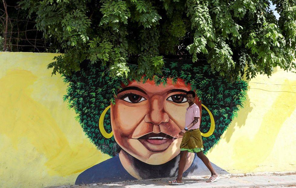 A boy walks past a wall with a woman's face painted on it. She has a green afro which blends into the trees growing behind the wall, giving the illusion that the trees are part of the artwork.