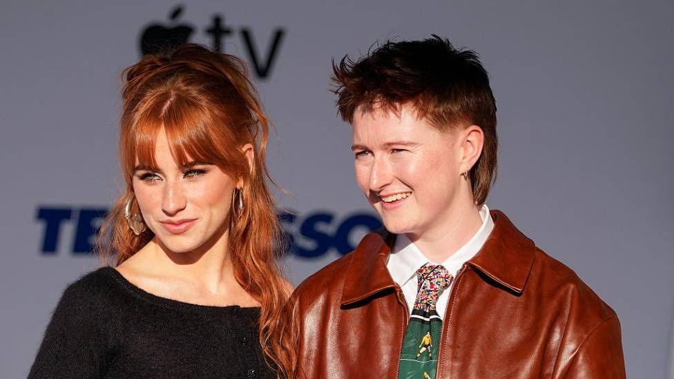 Two people standing side by side. One has long red hair and is wearing a black top. The other has short hair and is wearing a red jacket and green tie over a white shirt