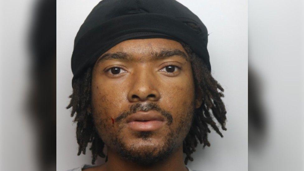 Mugshot of Khalil Muhammad, looking at the camera has dark hair and eyes, stubble, dreadlocked hair and is wearing a black hat
