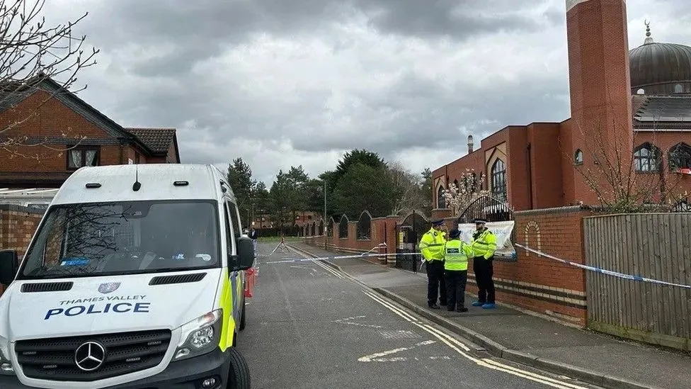 The same street scene in Manzil Way, but with a police van on the left of the picture and three police officers in front of the mosque on the right side of the road and the picture 